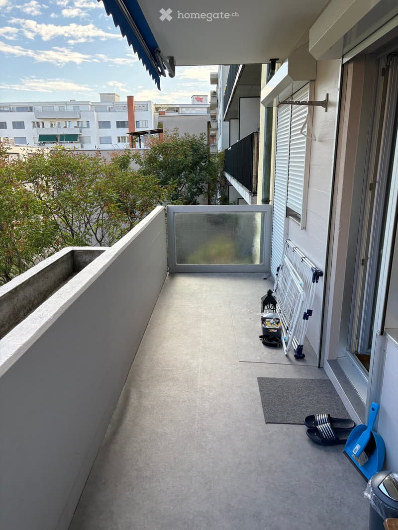 Balcony with concrete floor, white walls, and a view of surrounding buildings and greenery. Includes a small rug, slippers, a broom, and a folding chair.