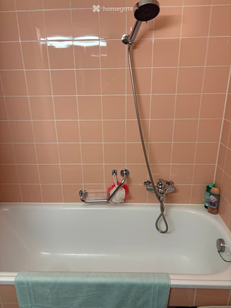 Bathroom with a white bathtub, pink tiled walls, a showerhead with a hose, a towel on the edge of the tub, and a few bathroom accessories on the side.