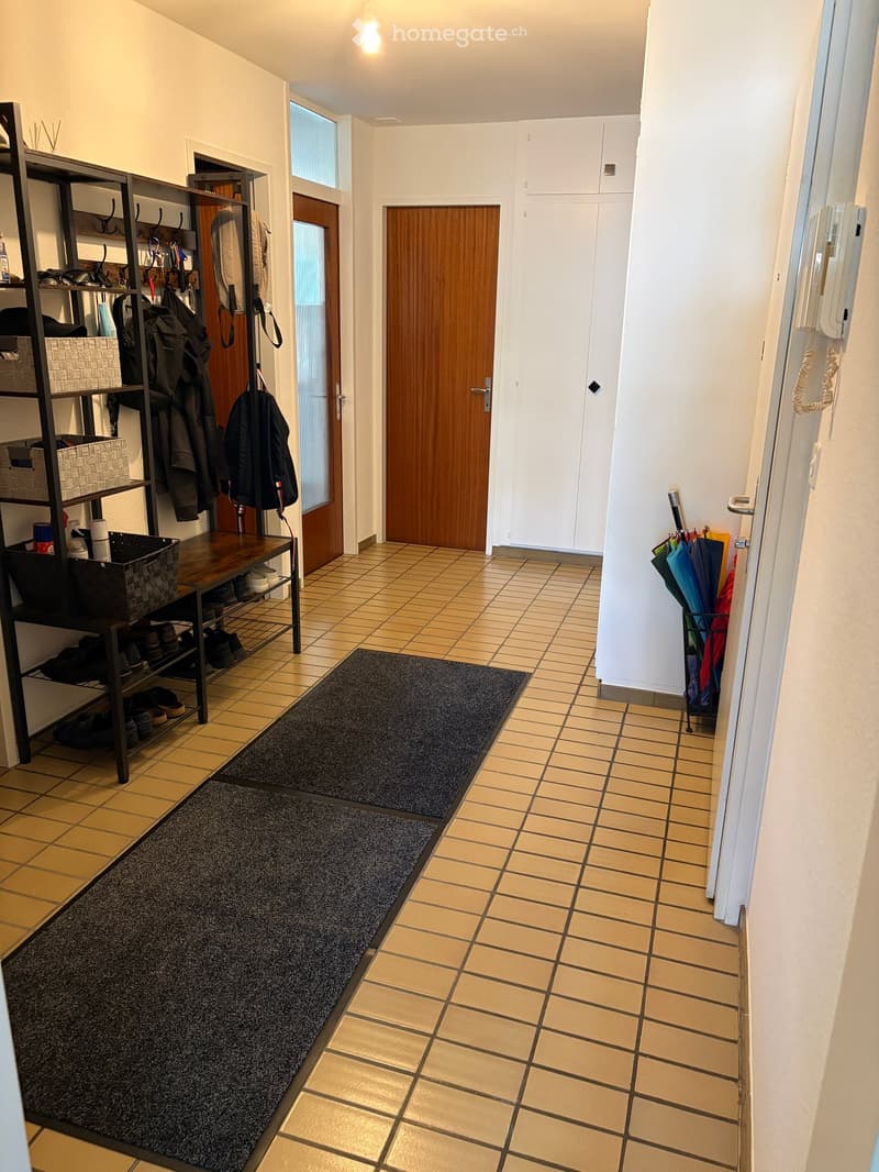 Hallway with tiled floor, white walls, coat rack, umbrella stand, and two black rugs leading to a wooden door and white closet.