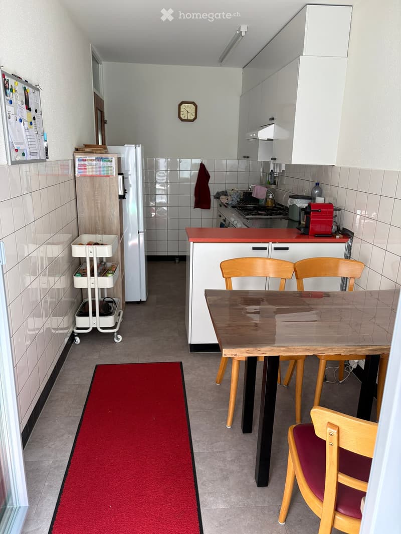A compact kitchen with white cabinets, a red countertop, a wooden dining table with chairs, a refrigerator, and a small cart with storage bins.