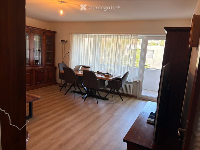 Brightly lit room with wooden flooring, a large window with white curtains, a wooden table with four chairs, a laptop, and a cabinet with glass doors.