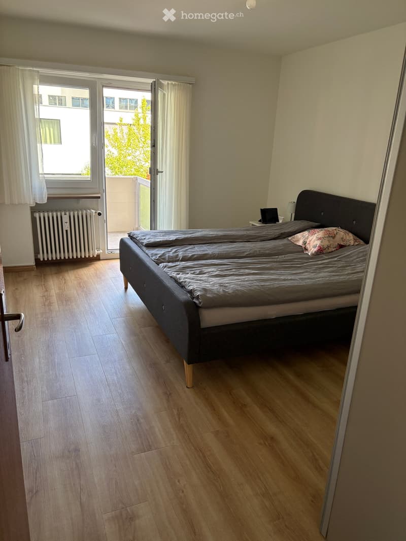 Bedroom with wooden flooring, double bed with dark headboard, nightstand, and lamp. Large window with white curtains and sliding glass door leading to a balcony.