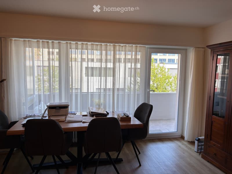 Brightly lit room with a wooden desk, two chairs, a laptop, printer, and a wooden cabinet near a large window with sheer curtains.