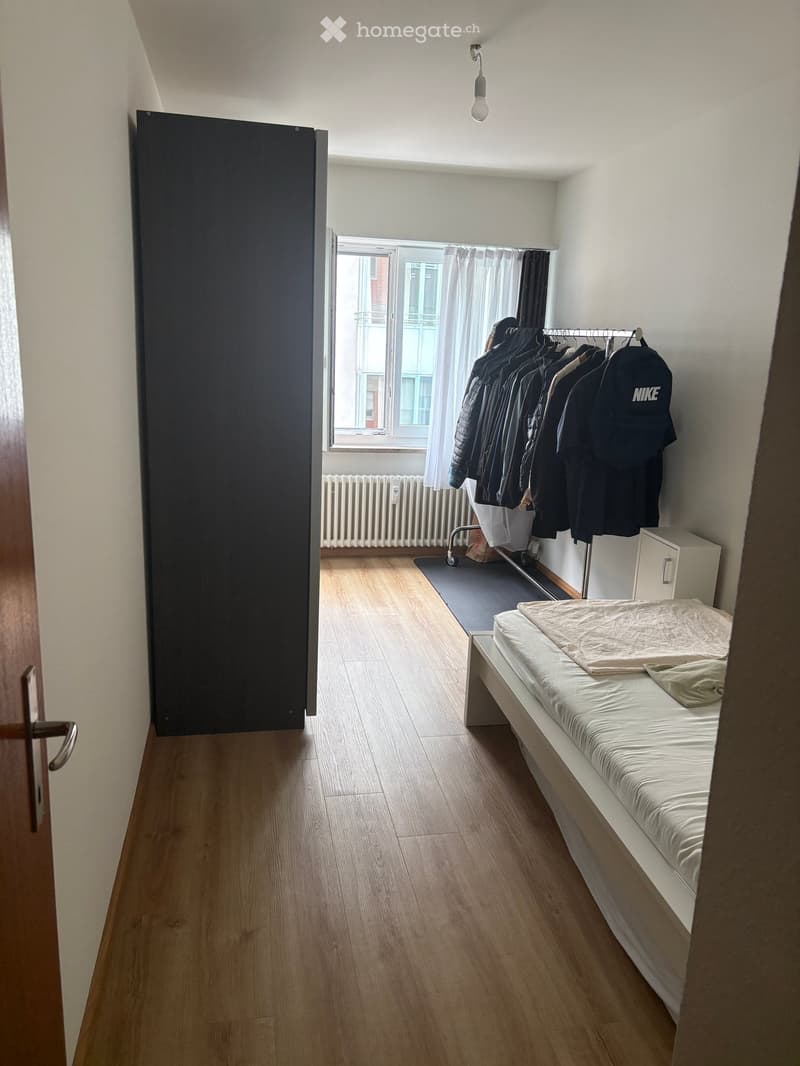 Small bedroom with wooden flooring, a single bed with white bedding, a clothing rack with jackets and shirts, a radiator under the window, and a partially open door.