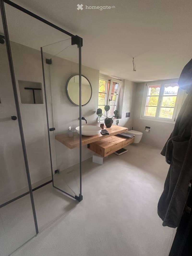 Modern bathroom with a wooden vanity, white sink, round mirror, and a glass-enclosed shower area.
