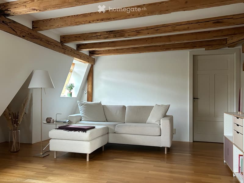 Modern living room with exposed wooden beams, light-colored sofa, ottoman, small table, floor lamp, and decorative vase.