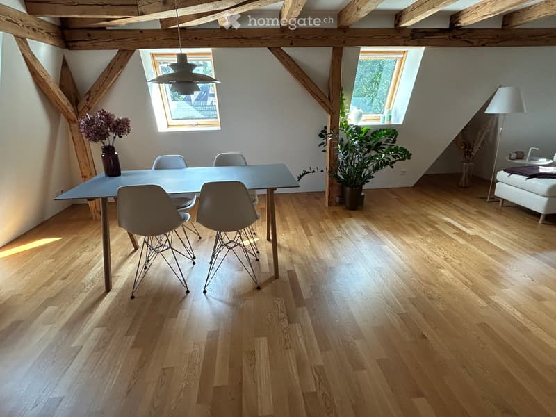 Bright, airy dining area with wooden floors, exposed wooden beams, and large windows. Features a modern dining table with four chairs, a vase with flowers, a potted plant, a sofa, and floor and pendant lamps.