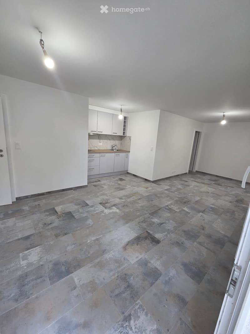 Empty room with grey tiled floor, white walls, and a small kitchen area with white cabinets and a sink.