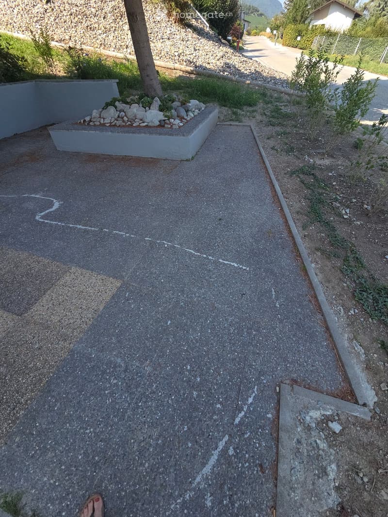 A narrow concrete pathway bordered by low stone walls, with small landscaped areas featuring rocks and sparse vegetation on either side.