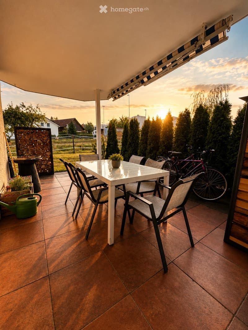 Balcony with a white table and chairs, bicycles, potted plants, and a garden hose, overlooking a suburban area at sunset.