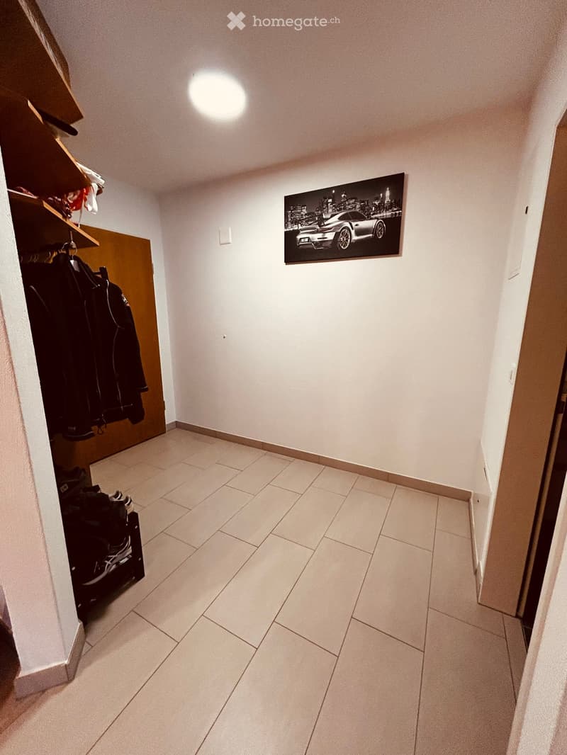 A clean, well-lit hallway with tiled flooring, white walls, and a large framed picture of a car on one wall. A coat rack with jackets and shoes is visible on the left side.