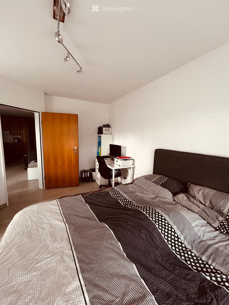 Modern bedroom with a large bed, patterned bedding, a desk with a computer, and a chair. The room features white walls and a wooden door leading to another area.