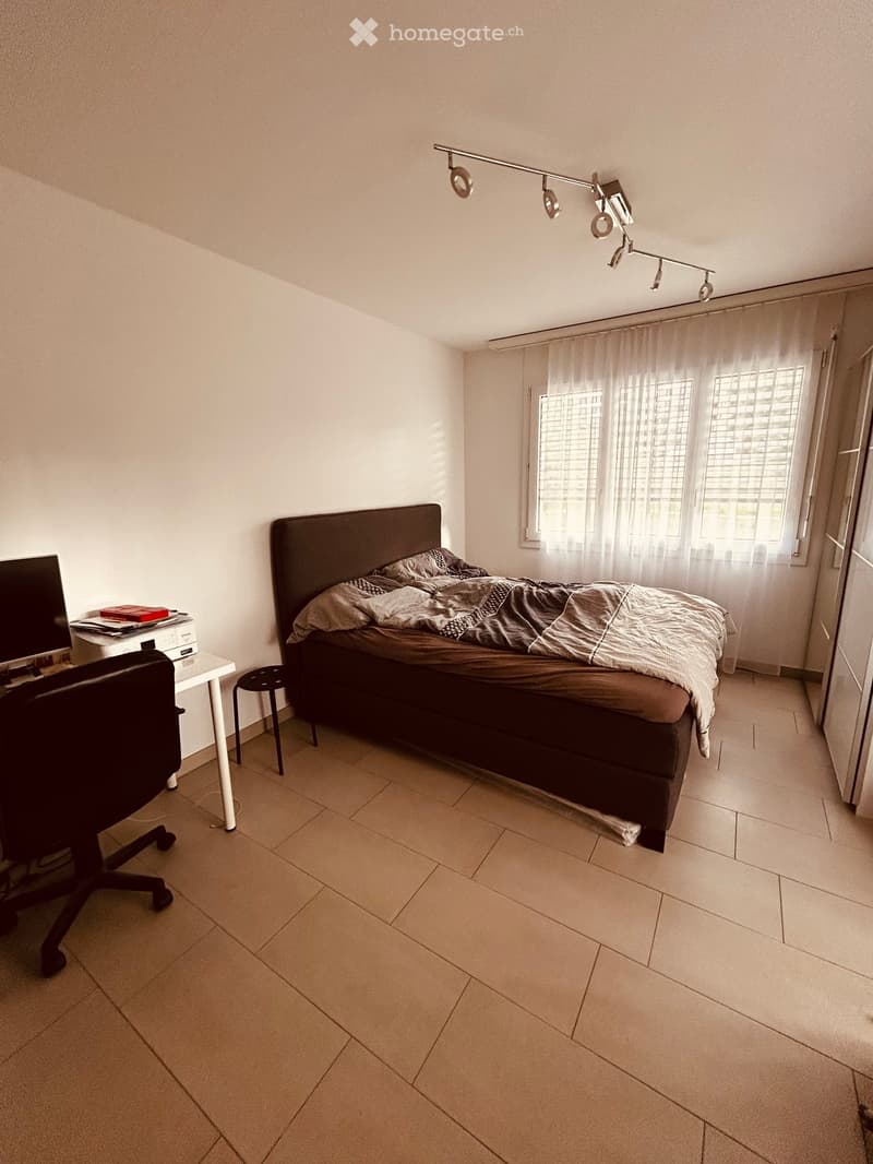 A modern bedroom with a large bed, a desk with a screen, a chair, and a stool. The room features tiled flooring, large windows with curtains, and a track light fixture above the bed.