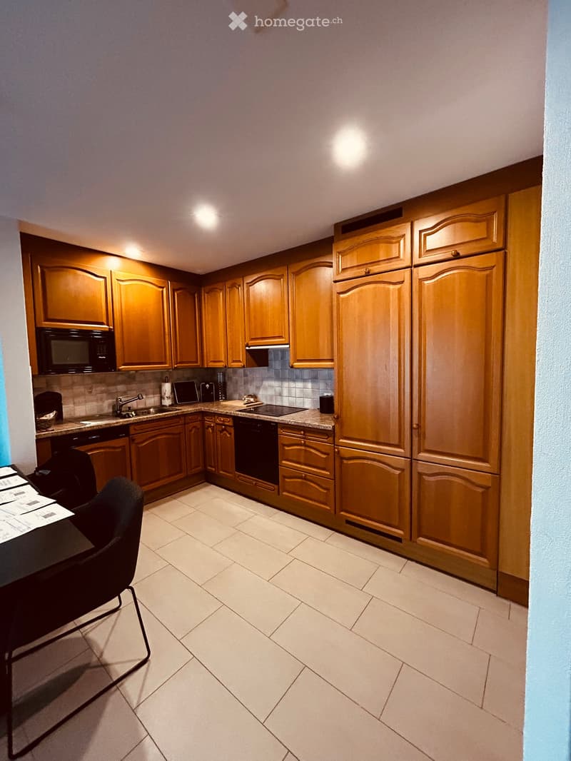A modern kitchen with wooden cabinets, stainless steel appliances, and a tiled floor. Includes a refrigerator, microwave, dishwasher, sink, and a small table with a chair.