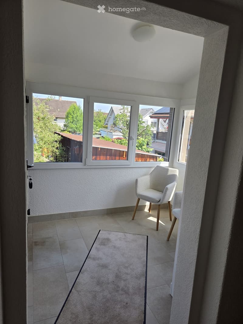 Balcony with tiled floor, white walls, large windows, and a single white chair with wooden legs.