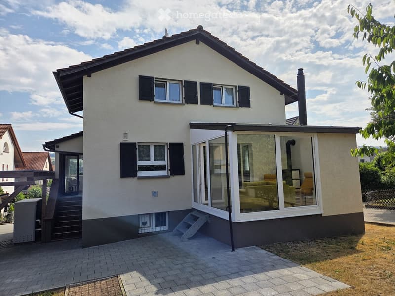 Two-story house with white exterior, black shutters, and a modern sunroom extension with large glass windows. Features a paved entrance area and steps leading to the main entrance.