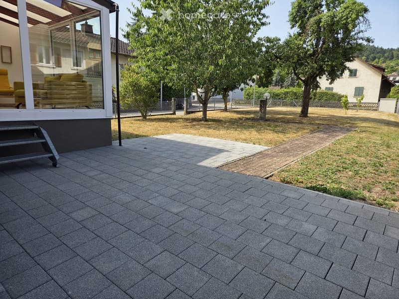 Garden area with a paved patio, steps leading to a house, trees, and a lawn.