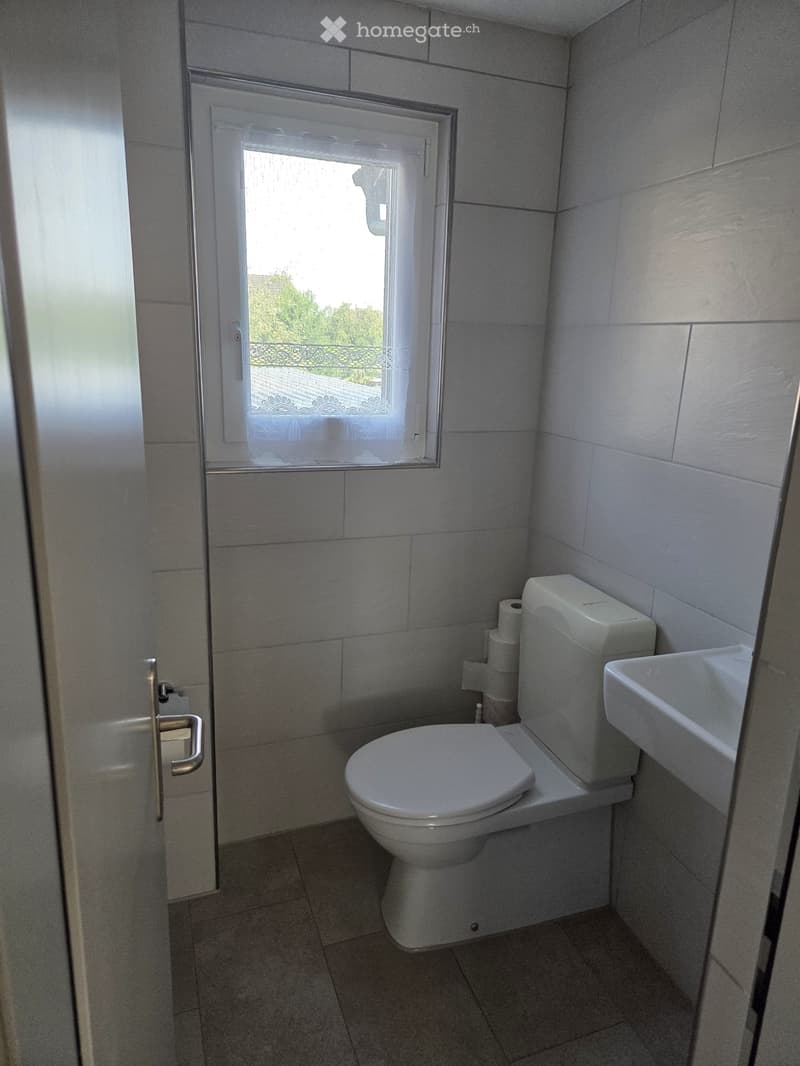 Small bathroom with white tiled walls and floor, featuring a toilet, a sink, and a window with a view of greenery outside.