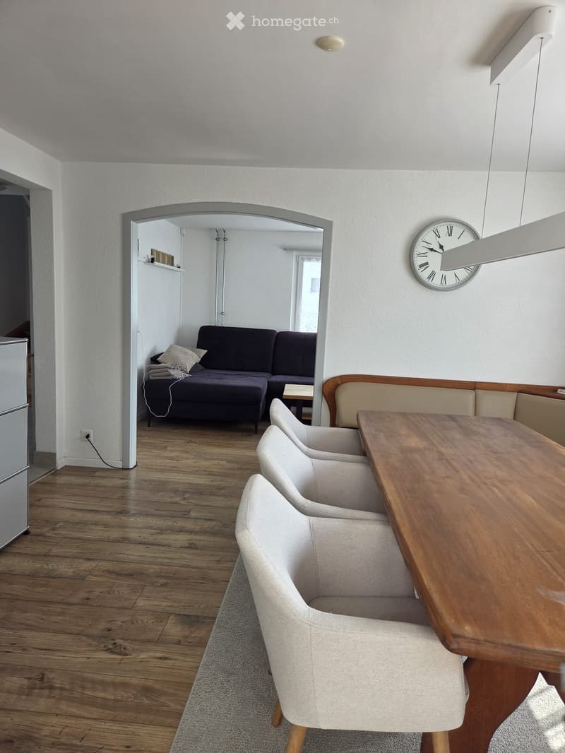 Modern living room with wooden flooring, a sofa, a dining table with chairs, and a clock on the wall.