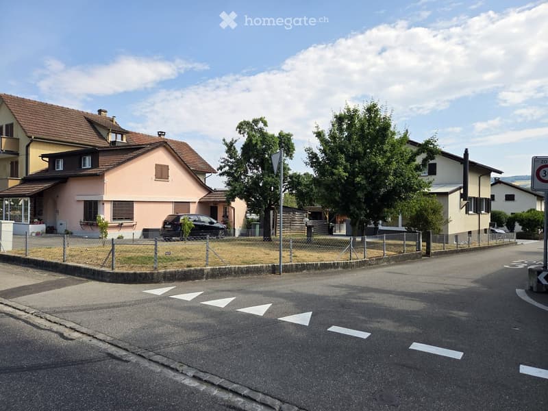 Residential street with multiple houses, including a pink house with a brown roof, a yellow house, and a white house. There is a parked car, a fence, and some greenery.