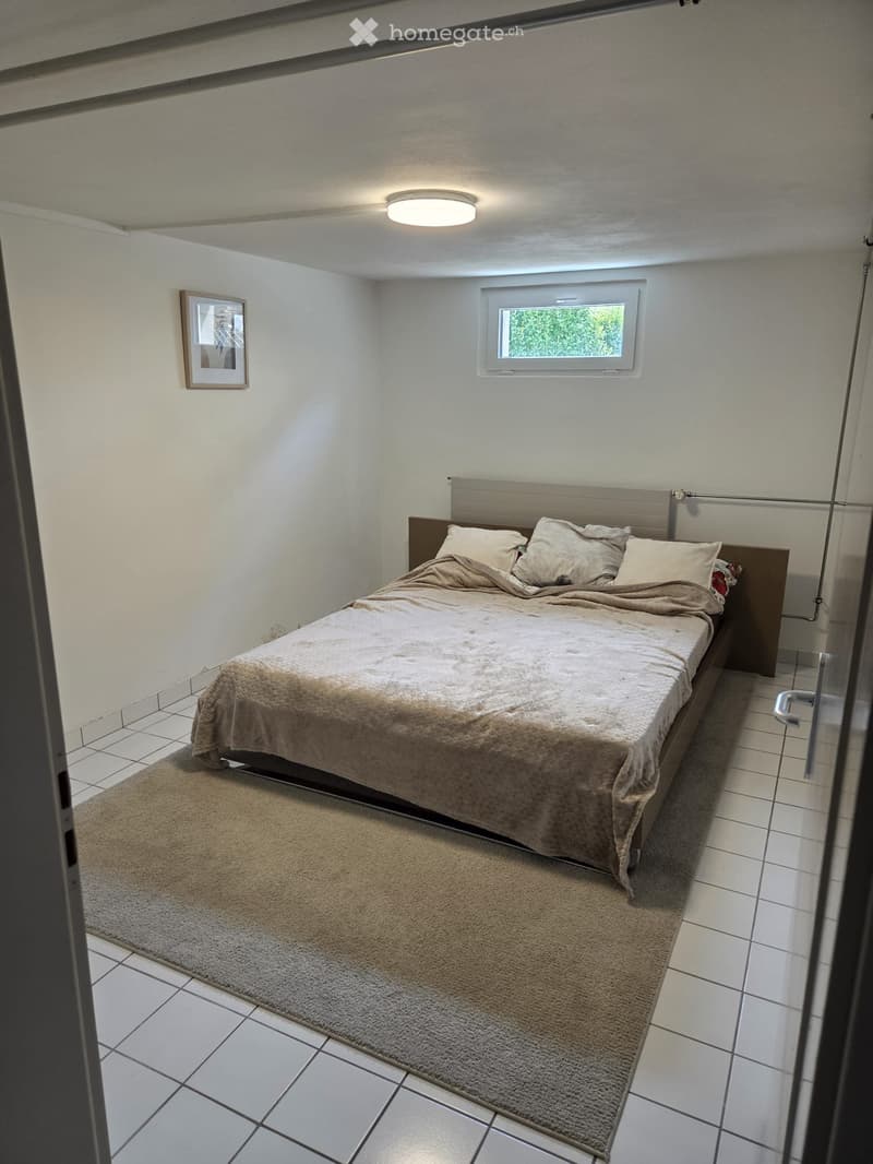 Cozy bedroom with a bed, pillows, blanket, and a small rug on the floor. A picture frame is on the wall, and a window provides natural light.