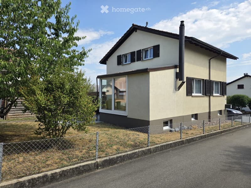 Two-story house with beige exterior, dark roof, multiple windows with shutters, and a fenced yard.