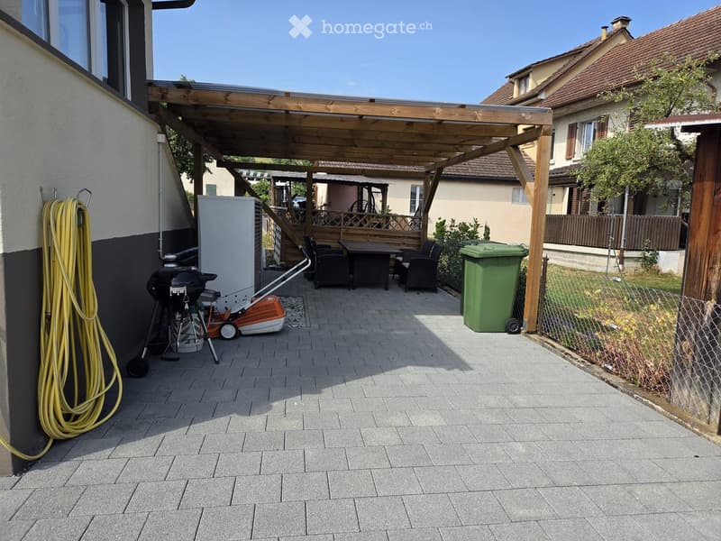 Patio area with a wooden pergola, seating area with a table and chairs, barbecue, lawn mower, refrigerator, and trash bin.
