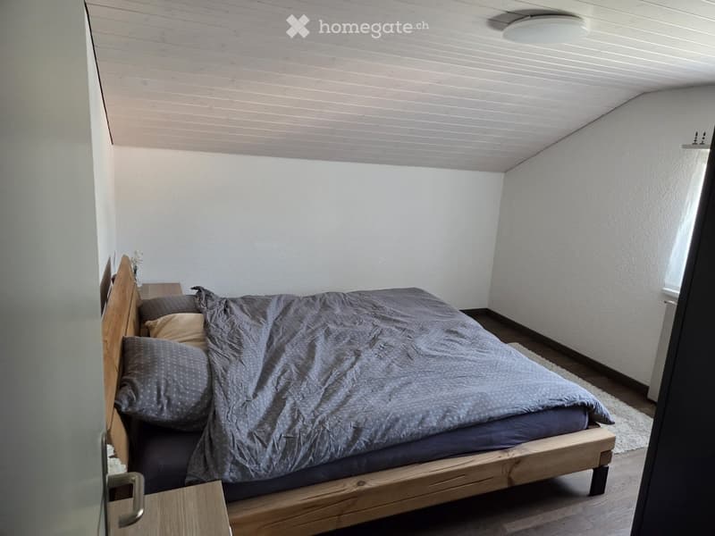 Cozy bedroom with a wooden bed frame, grey bedding, and a nightstand with a vase of flowers. The room features light-colored walls and a slanted wooden ceiling.