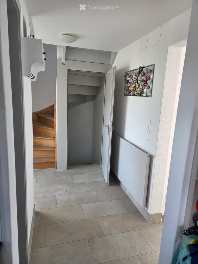 A narrow hallway with tiled flooring, white walls, and a staircase leading upwards. A radiator is mounted on the right wall, and a small shelf with various items is visible near the staircase.