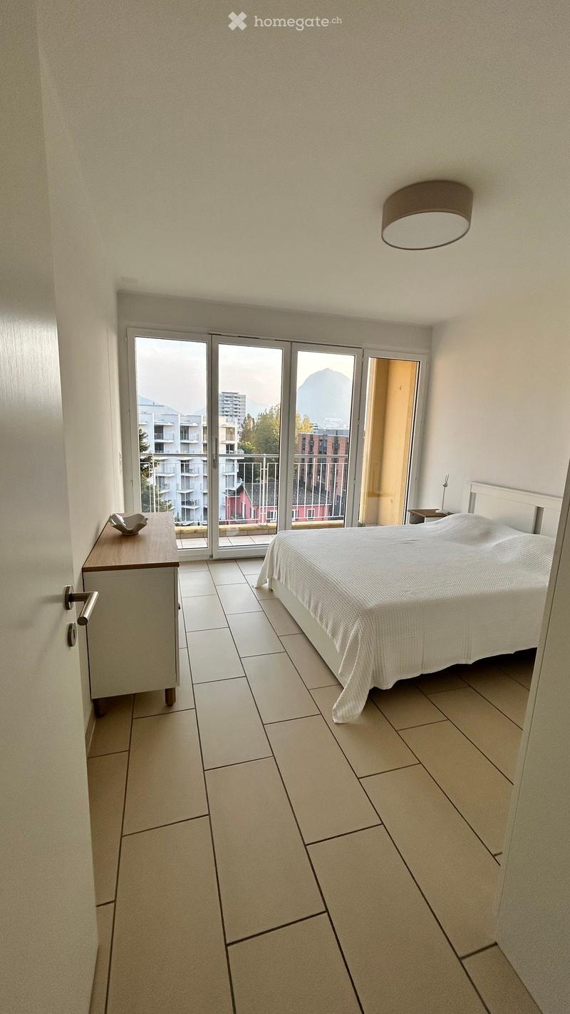 Modern bedroom with a large bed, nightstand, small table, and a balcony with a scenic mountain view.