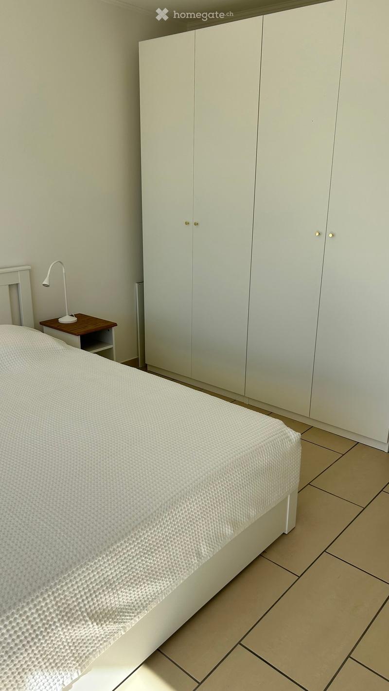 A modern bedroom with a white bed, a nightstand with a lamp, and a large white wardrobe.