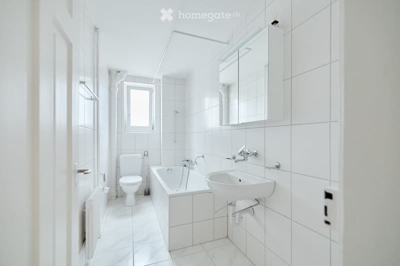 Modern, white-tiled bathroom with a toilet, bathtub, sink, mirror, and cabinet.