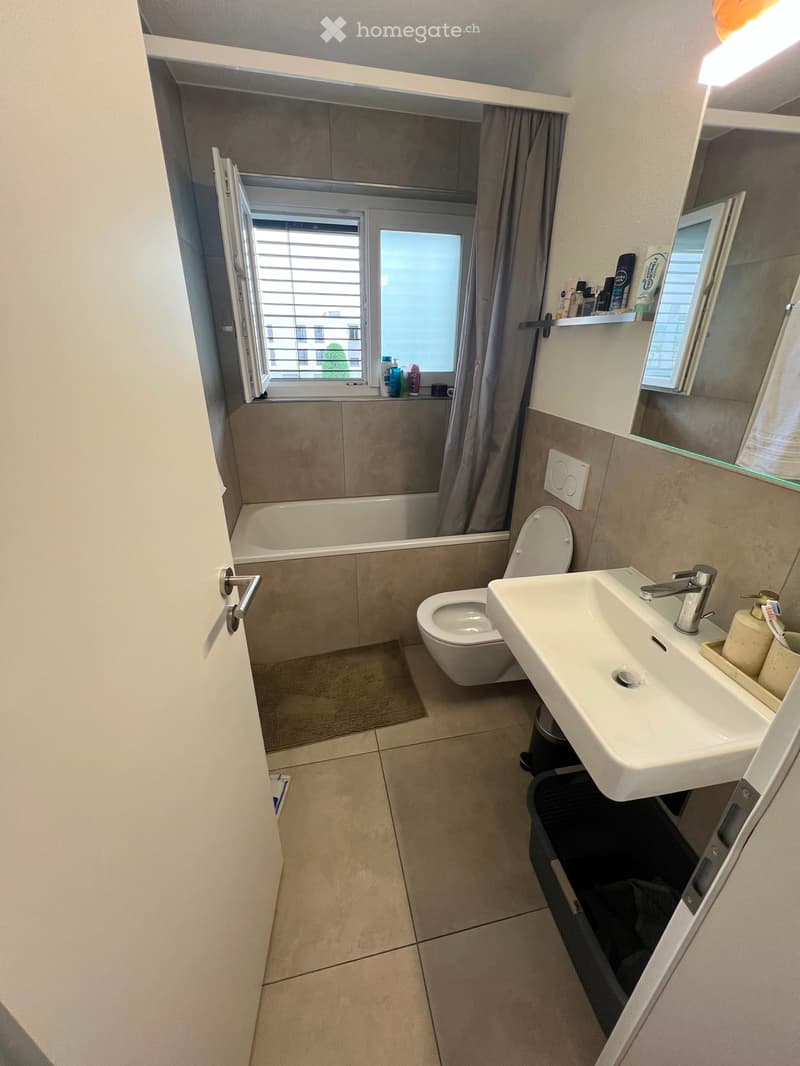 Modern bathroom with a white sink, toilet, bathtub, and mirror. The room features tiled walls and floor, a window with blinds, and a small shelf with toiletries.
