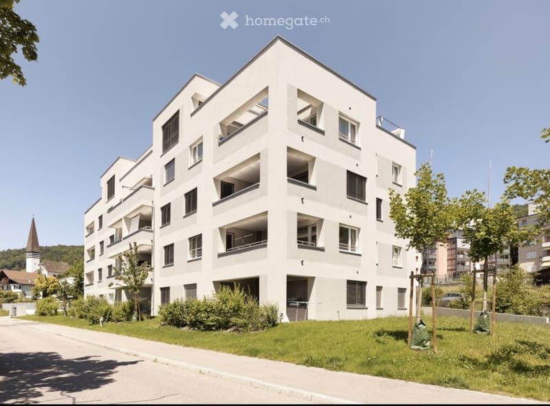 Modern multi-story residential building with white facade, numerous windows, and balconies. Surrounded by greenery and a paved pathway.