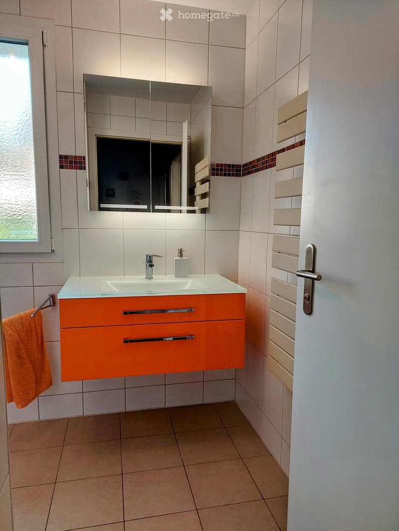 Modern bathroom with white tiled walls, a glass sink top, orange cabinet, and a large mirror above the sink.