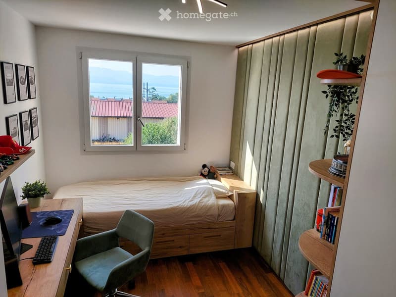 Cozy bedroom with a single bed, desk, chair, and shelves. Features a large window with a scenic view, framed pictures on the wall, and a small plant on the desk.