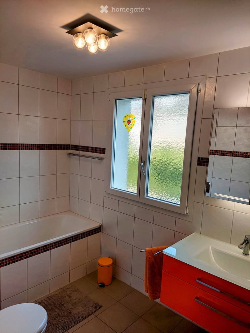 Modern bathroom with white tiles, a red cabinet, a white sink with a chrome faucet, a mirror, a bathtub, a toilet, and an orange trash can. Features a frosted glass window with a heart design and a towel hanging on a rack.