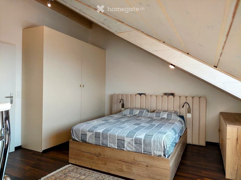 Modern bedroom with a wooden bed, upholstered headboard, patterned bedding, and a small nightstand with books. The room features wooden flooring and built-in wardrobes.