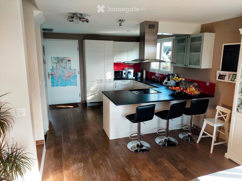 Modern kitchen with white cabinets, black countertop, and stainless steel appliances. Features a central island with four black bar stools, a refrigerator, microwave, and coffee maker on the counter.