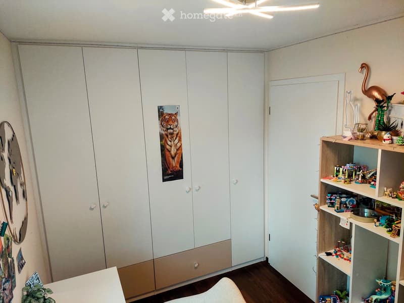 A well-lit room with a large white wardrobe, a wooden shelf filled with toys and decorative items, a small table, and a plant on the floor.