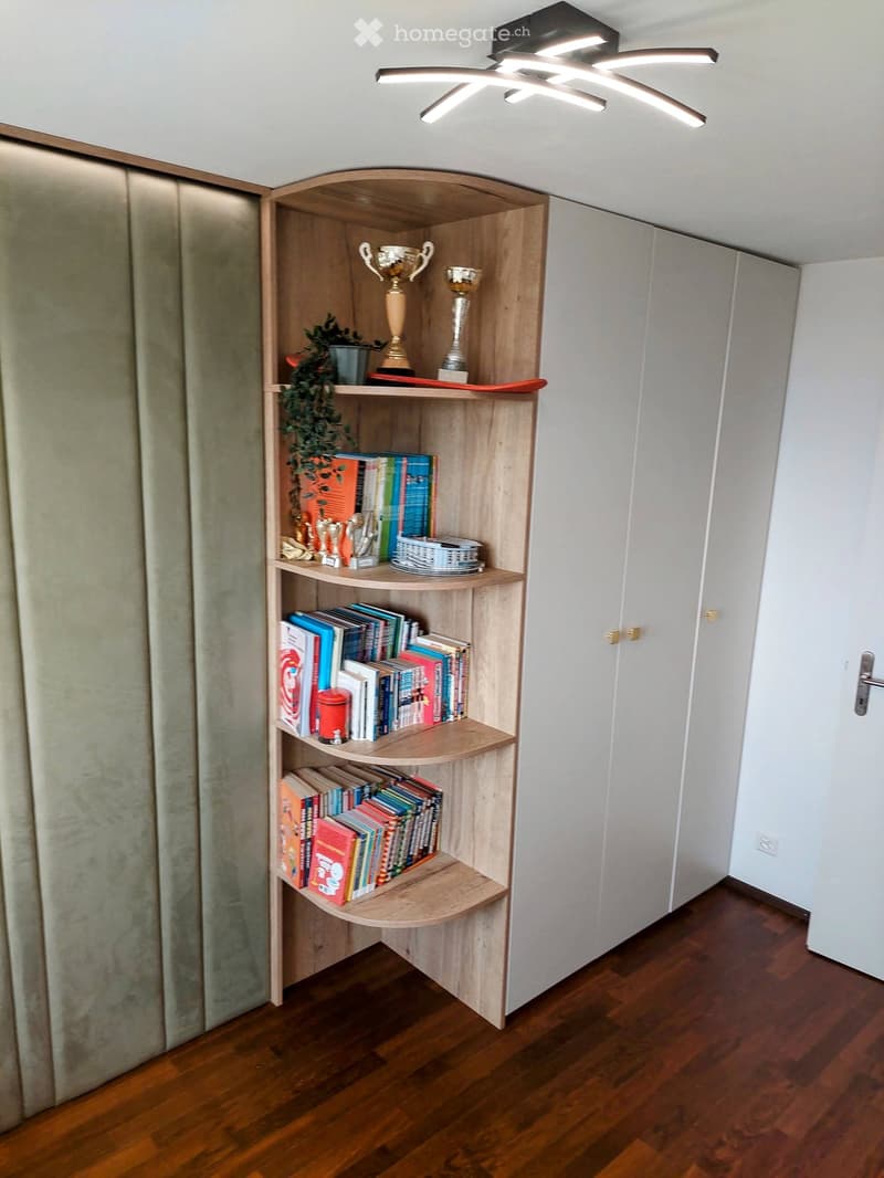 A wooden bookshelf filled with books, trophies, and a small plant, positioned against a white wall with a modern ceiling light fixture above.