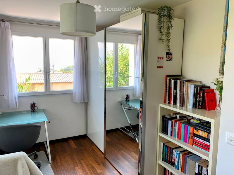 Well-lit room with wooden flooring, large windows, a modern desk with a chair, a bookshelf filled with books, and a hanging plant. A mirror and pendant light are also visible.