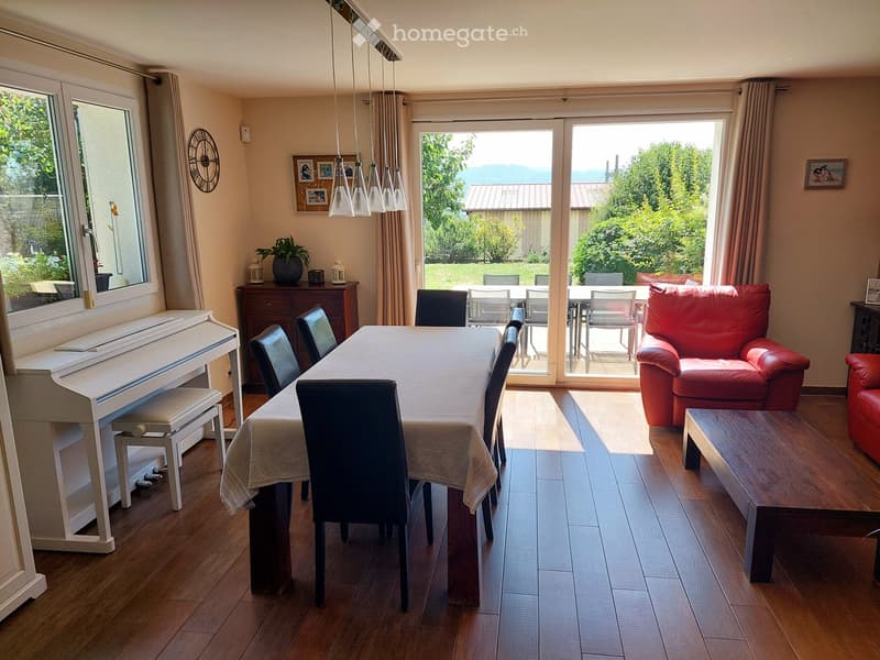 A well-lit living and dining area with wooden flooring, a red leather sofa, a dining table with chairs, a white piano, and a view of a garden through large windows and sliding doors.