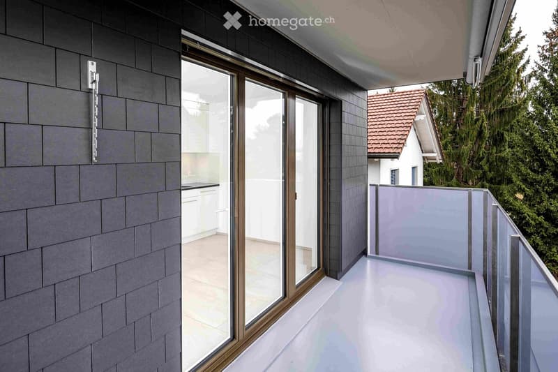 Modern balcony with sleek glass railing, white flooring, and a sliding glass door leading to an interior space with white cabinets and a countertop.