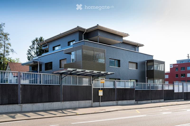 Modern two-story building with grey brick facade, large windows, and a flat roof. Features a covered entrance area with a metal fence and a paved parking area.
