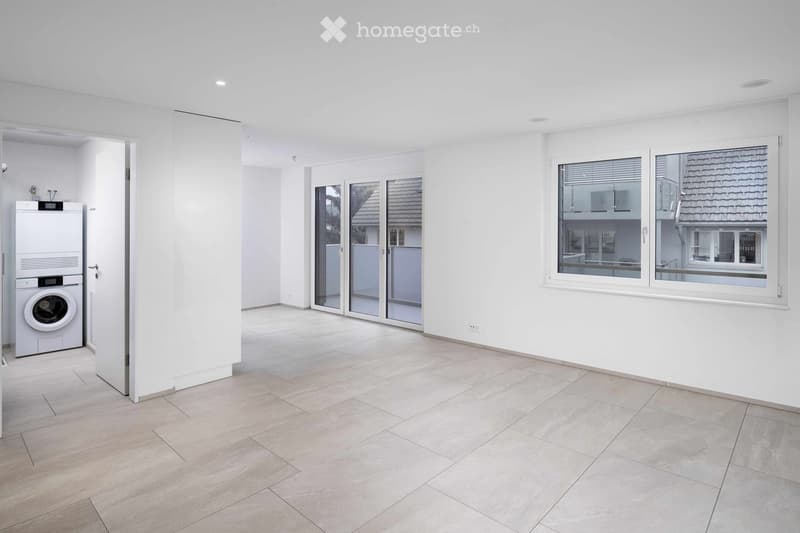 Empty room with tiled floor, white walls, large windows, and a private washing machine in the corner.