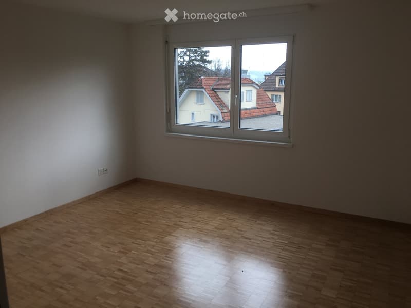 Empty room with wooden flooring and large window showing a view of neighboring houses.