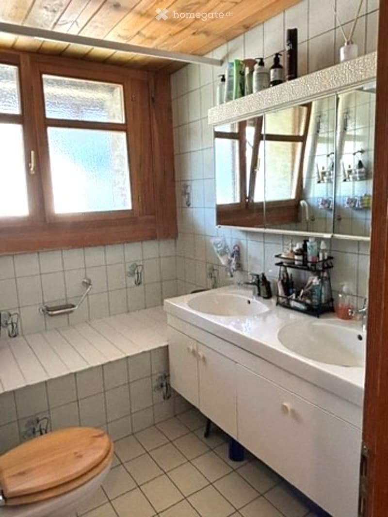 Bathroom with white tiled walls and floor, double sink vanity with mirrored cabinet, toilet with wooden seat, bathtub, and shower area.