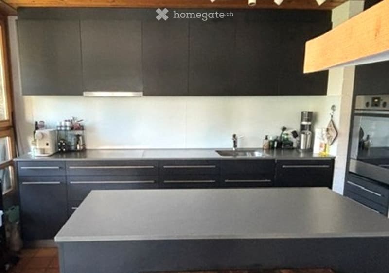 Modern kitchen with dark grey cabinets, a central island, and stainless steel appliances including a sink, oven, and fridge. A coffee maker and other small appliances are on the counter.