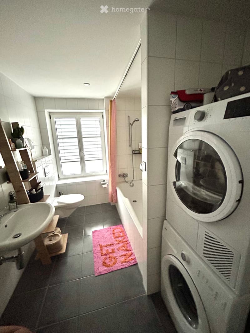 Bathroom with a sink, toilet, bathtub, and a washing machine. Features a pink bath mat and a window with blinds.
